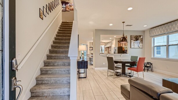 A modern interior with a staircase leading to a dining area, a kitchen in the background, and a large window providing natural light. - Sunset Home with Modern Living at Encore (Kissimmee)