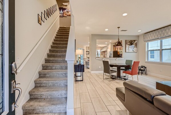A modern interior with a staircase leading to a dining area, a kitchen in the background, and a large window providing natural light.