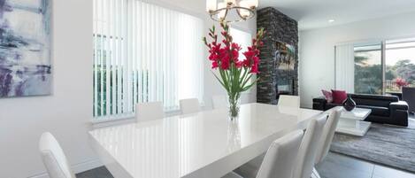 A modern dining room with a white table, chairs, and a vase of red flowers. There is a stone fireplace and a black sofa in the background.