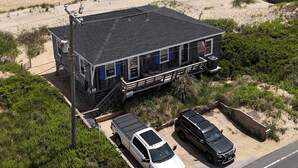 Ein Anwesen am Strand mit einem Haus, einem Balkon, einer Einfahrt mit zwei Autos und Blick auf den Ozean.