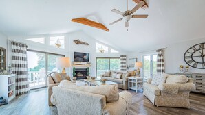 A spacious living room with a fireplace, comfortable seating, and large windows offering a view of the outdoors. - Captain's Perch - Duck, Northpoint, 400 Yds to Beach, Pool, Hot Tub, Events, Flex Stays (Duck)