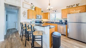A kitchen with wooden cabinets, a stainless steel refrigerator, a microwave, and a sink. There is a bar area with stools and a trash can. - Captain's Perch - Duck, Northpoint, 400 Yds to Beach, Pool, Hot Tub, Events, Flex Stays (Duck)