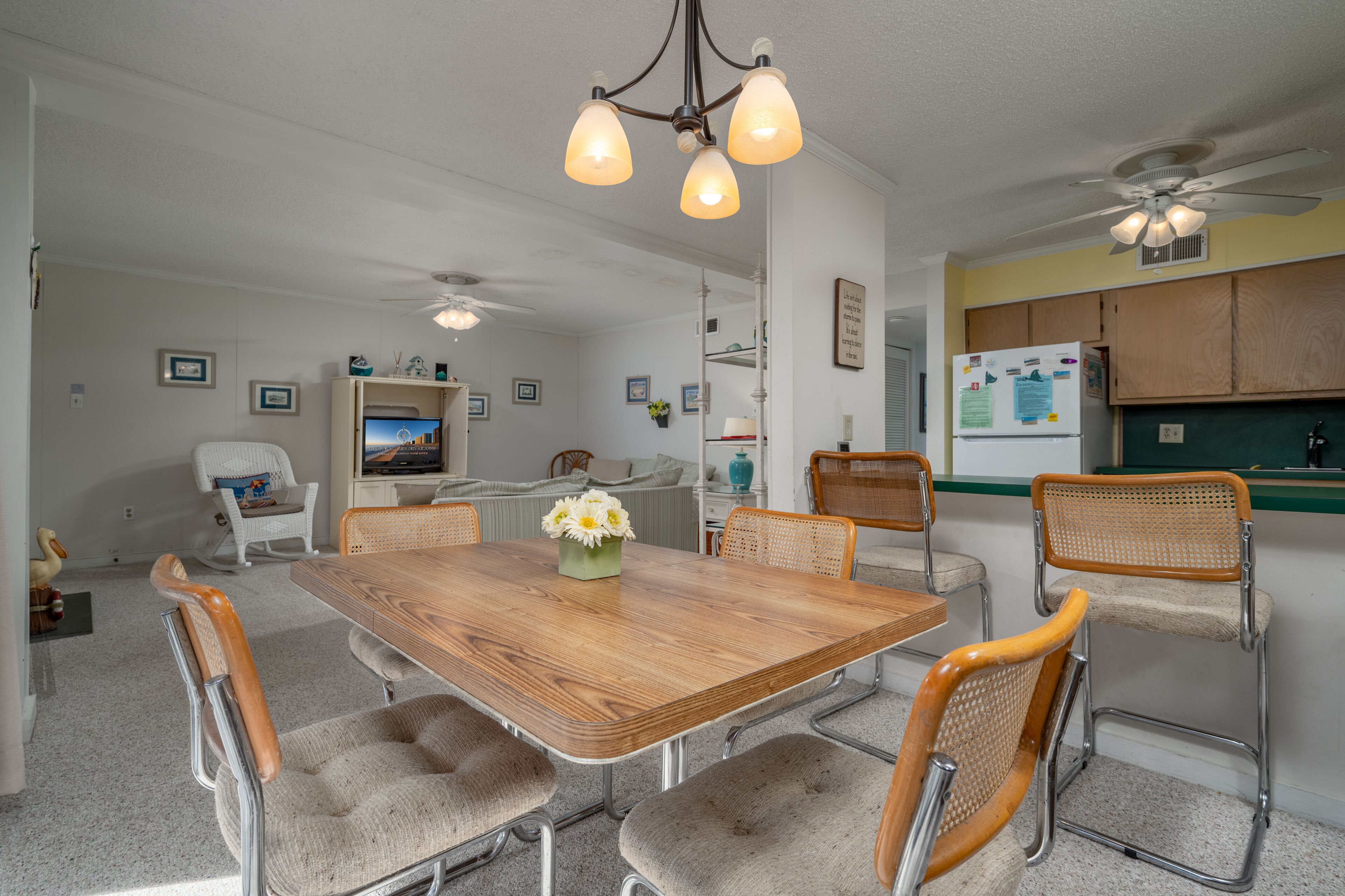 A dining area with a wooden table, six upholstered chairs, a chandelier, ceiling fans, and a kitchen area with wooden cabinets.
