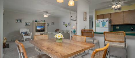 A dining area with a wooden table, six upholstered chairs, a chandelier, ceiling fans, and a kitchen area with wooden cabinets.