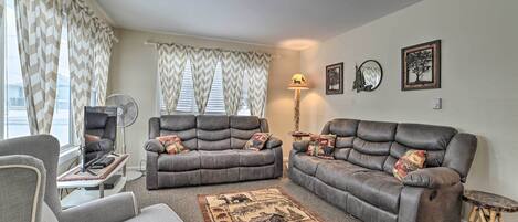 Uma sala de estar com dois sofás cinzas, uma poltrona cinza, uma mesinha, um abajur e um tapete decorativo.