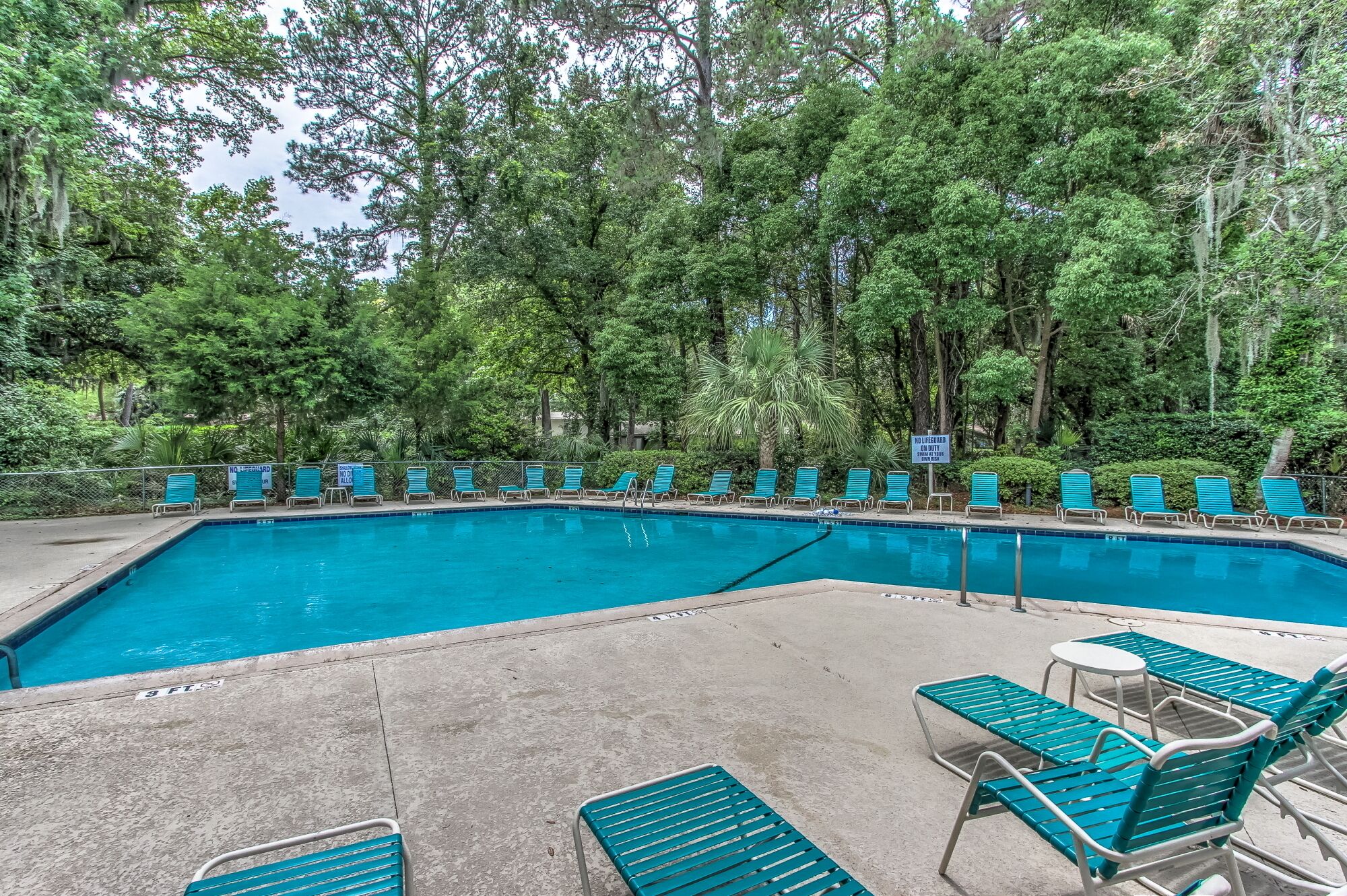 Una piscina con agua azul, rodeada de árboles y sillas de descanso verdes.