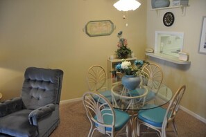 A dining area with a glass table, four chairs, a grey armchair, a vase with flowers, a clock, and a framed picture on the wall.