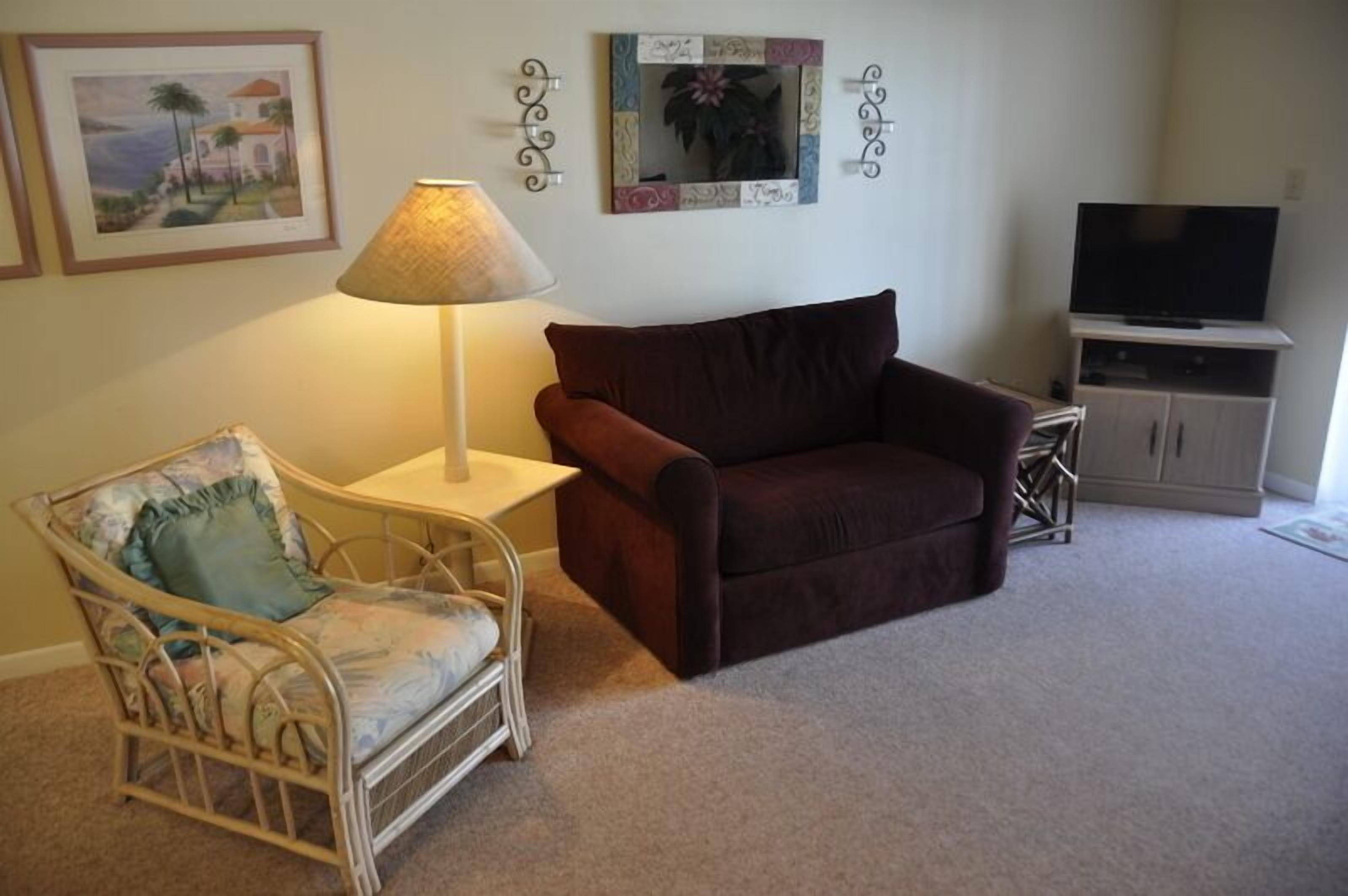 A living room with a brown sofa, a wicker chair with a cushion, a lamp, a television on a stand, and framed pictures on the wall.