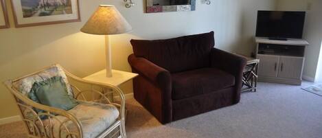 A living room with a brown sofa, a wicker chair with a cushion, a lamp, a television on a stand, and framed pictures on the wall.