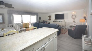 Ein geräumiges Wohnzimmer mit großem Fenster, ein Essbereich mit weißen Stühlen, eine Küche mit Marmorarbeitsplatte und ein dunkel gefärbter Sessel.