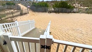 Blick auf einen Strand von einer Terrasse mit weißer Geländer, feinem Sand und einem Zaun im Hintergrund.