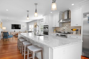 A modern kitchen with white cabinetry, a large island with bar stools, stainless steel appliances, and pendant lighting. - Historic Sea Captain's Home in Downtown Chatham (Chatham)
