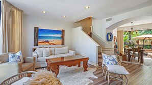 Un salón amplio con un sofá blanco, una mesa de centro de madera y sillas de mimbre. Un cuadro grande con una escena de playa decora la pared.