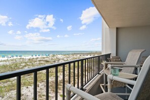 A balcony with wicker chairs and a glass table overlooking a beach with umbrellas and a wooden walkway.