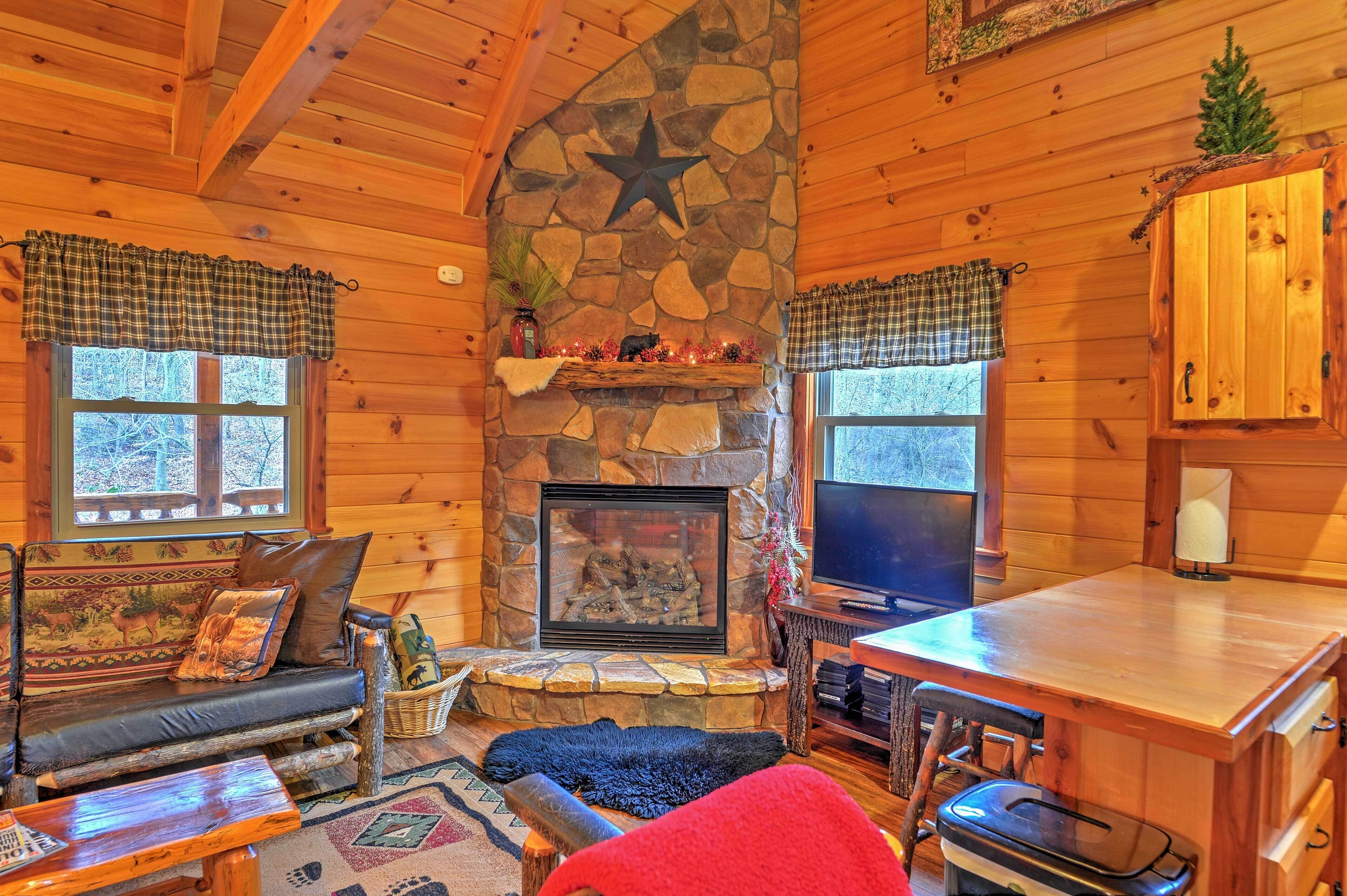 A cozy cabin interior with a stone fireplace, wooden walls, a sofa, a dining table, and a television.