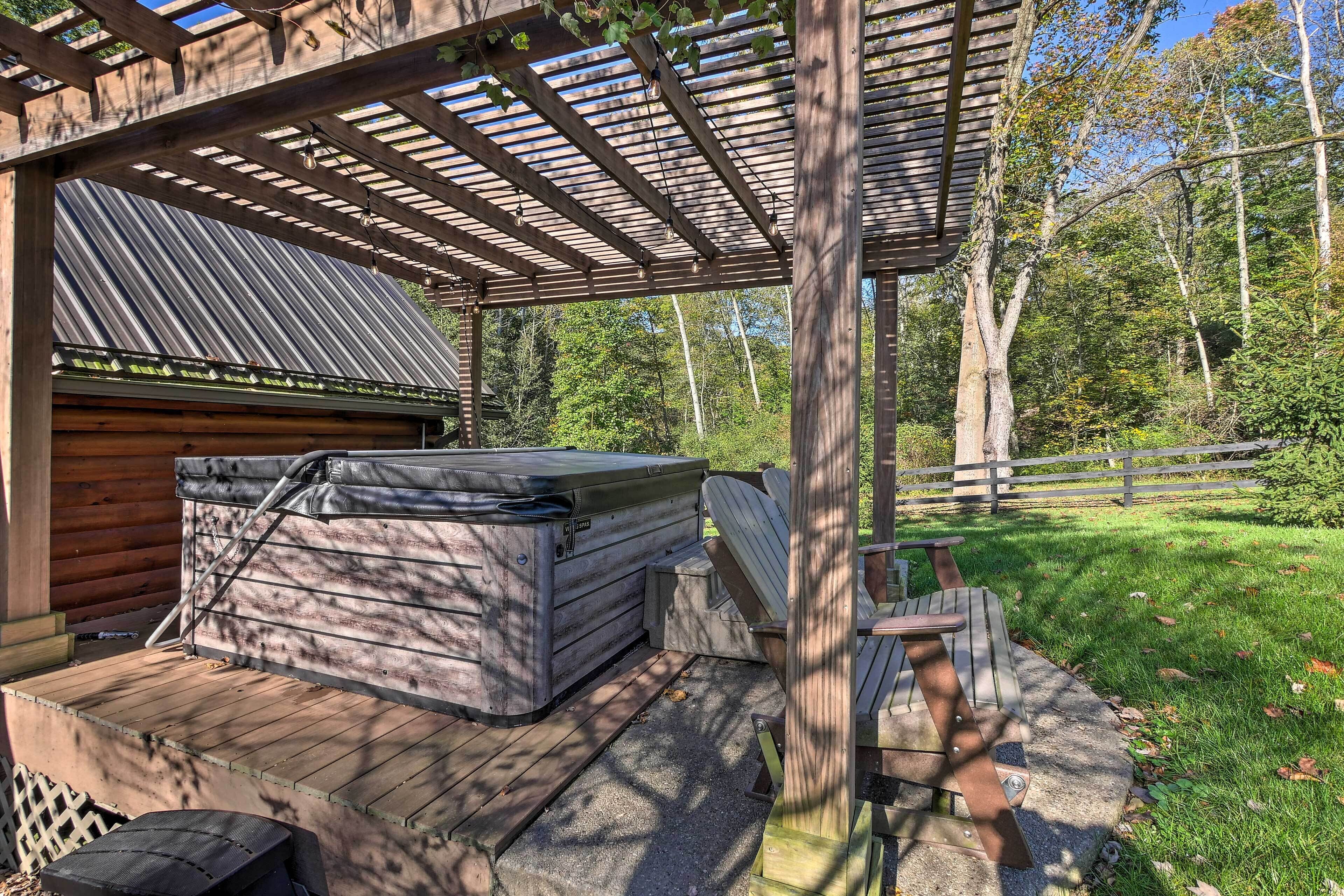 Una terraza de madera con un jacuzzi y un área de estar cubierta, rodeada por una valla de madera y árboles.