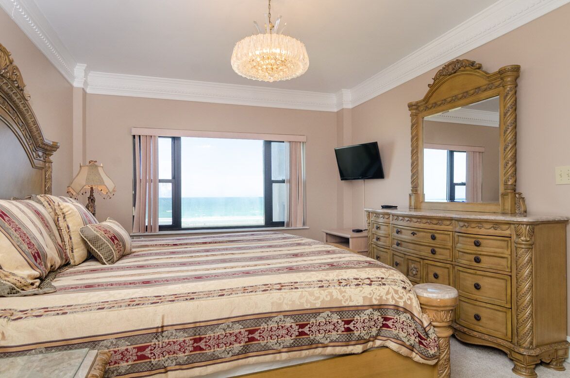 A bedroom with a large bed, a dresser, a television, and a view of the ocean.
