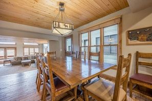 A spacious dining area with a wooden table and chairs, large windows, and a ceiling with wooden beams.