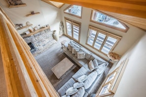 A cozy living room with a stone fireplace, a grey sofa with cushions, and multiple windows providing natural light.