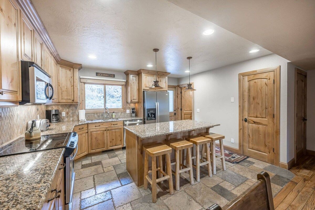 A kitchen with granite countertops, wooden cabinets, a central island with stools, and a refrigerator.
