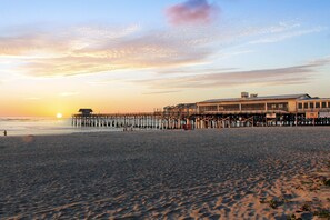 A beach with a pier extending into the water, a building with a flat roof, and the sun setting in the background.