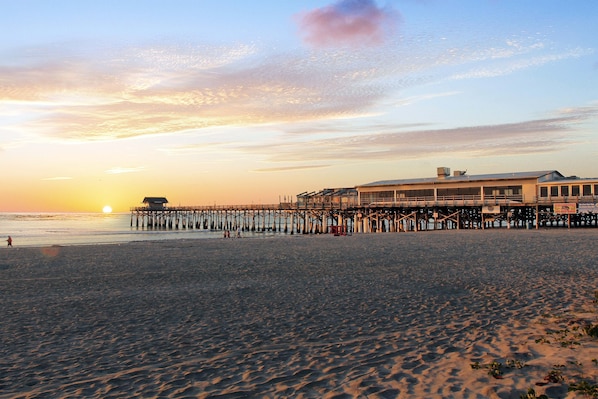 A beach with a pier extending into the water, a building with a flat roof, and the sun setting in the background.
