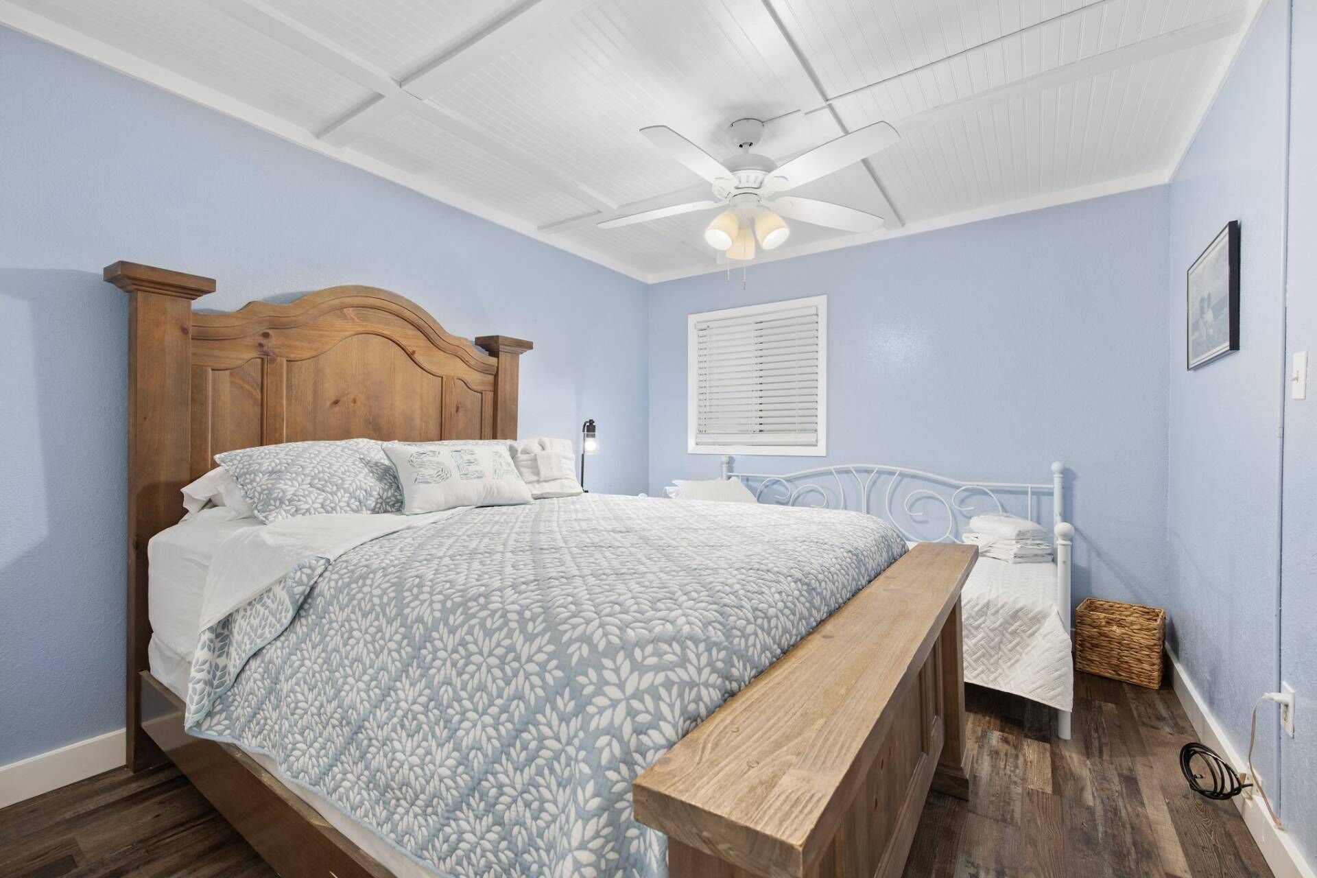 Une chambre à coucher avec un lit en bois, une table de chevet blanche, un ventilateur de plafond et une fenêtre avec des stores.
