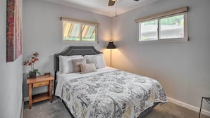 Un dormitorio con cama, mesita de noche, lámpara, ventilador de techo y dos ventanas.