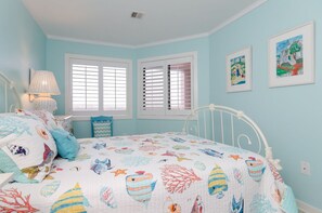Quarto com cama, duas janelas com persianas, uma luminária e duas obras de arte emolduradas na parede.