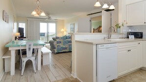 A modern kitchen with white cabinetry, a dishwasher, and a dining area with a glass table and white chairs. - Beautiful oceanfront condo in Ocean Dunes! (Wrightsville Beach)