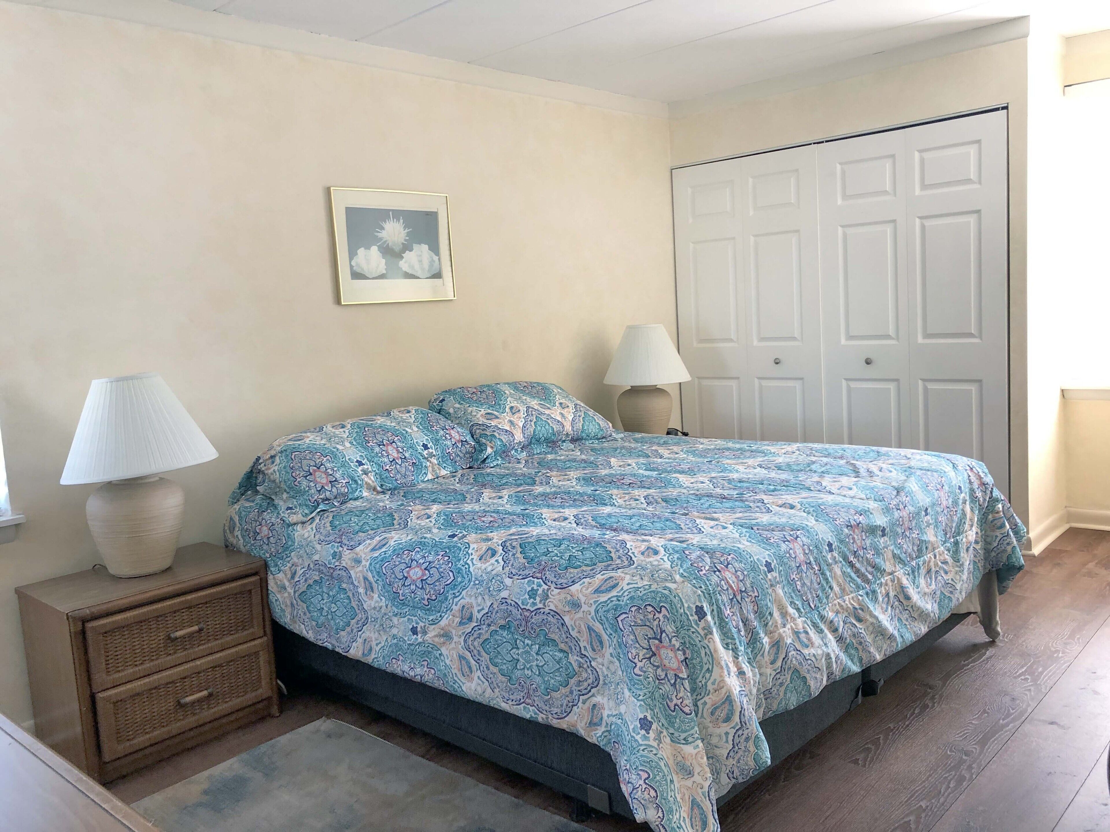 Une chambre à coucher avec un lit, deux tables de chevet avec des lampes, une porte et un tableau encadré au mur.