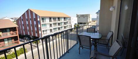 A balcony with a table and chairs, overlooking a street with buildings and a clear sky.