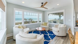 A spacious living room with large windows offering ocean views, a ceiling fan, and a blue patterned rug.