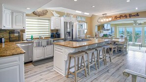 Una cocina amplia con armarios blancos, una isla grande con taburetes, un refrigerador de acero inoxidable y vistas al océano a través de puertas de vidrio.