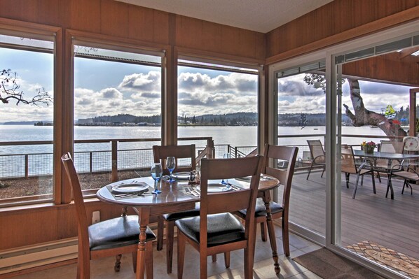 Comedor con mesa lista para cuatro personas, ventanas amplias que ofrecen vista a un lago y colinas al fondo.