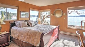 Une chambre à coucher avec un grand lit, une table de chevet, une commode et une chaise. Il y a deux fenêtres qui donnent sur un plan d’eau et sur des arbres.