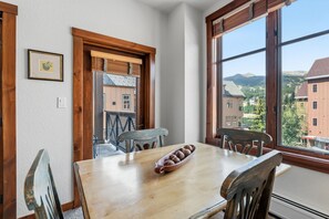 A dining area with a wooden table and chairs, a window with a view of buildings and mountains, and a door with a wooden frame.