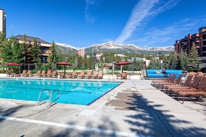 A pool area with lounge chairs and umbrellas, surrounded by buildings and mountains.