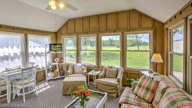 A cozy room with a ceiling fan, large windows with a view of a green landscape, a plaid sofa, and a white table with a vase of flowers. - Historic Ottawa Lake House: Deck, Barn + 96 Acres! (Ottawa Lake)