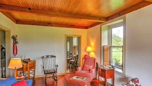 A cozy living room with a wooden ceiling, a rocking chair, a red checkered armchair, a table with a lamp, and a window with a view of greenery. - Historic Ottawa Lake House: Deck, Barn + 96 Acres! (Ottawa Lake)