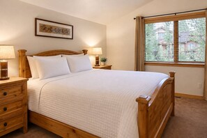 A bedroom with a wooden bed, two bedside tables with lamps, a window with blinds, and a framed picture on the wall.