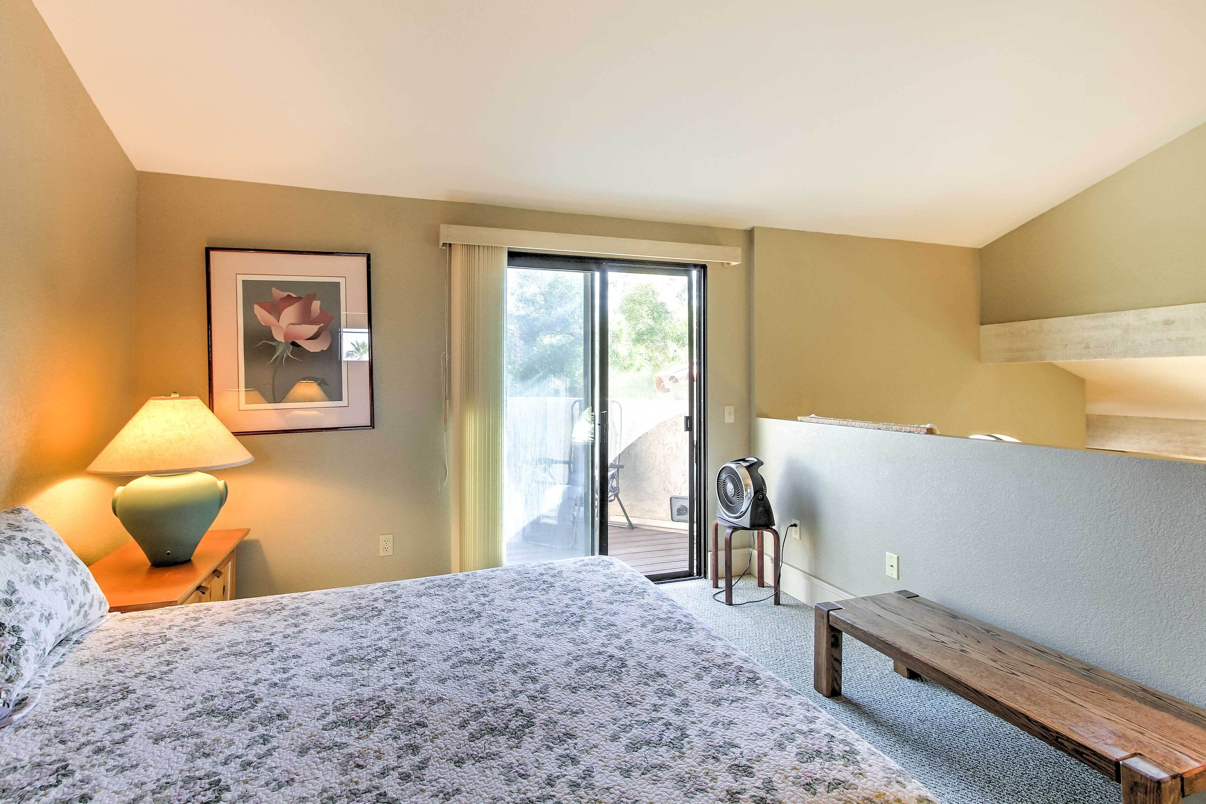 A hotel room with a bed, a desk with a lamp, a fan, and a sliding glass door leading to an outdoor area.