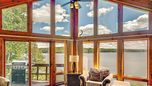 Une pièce spacieuse avec de grandes fenêtres donnant sur un lac et une forêt. À l’extérieur, on trouve une terrasse en bois, un barbecue et un coin salon confortable avec une lampe.