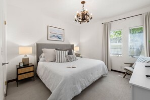 Ein Schlafzimmer mit einem großen Bett, zwei Nachttischen mit Lampen, einer Chandelüster, einem Fenster mit Jalousien und einem Sessel.