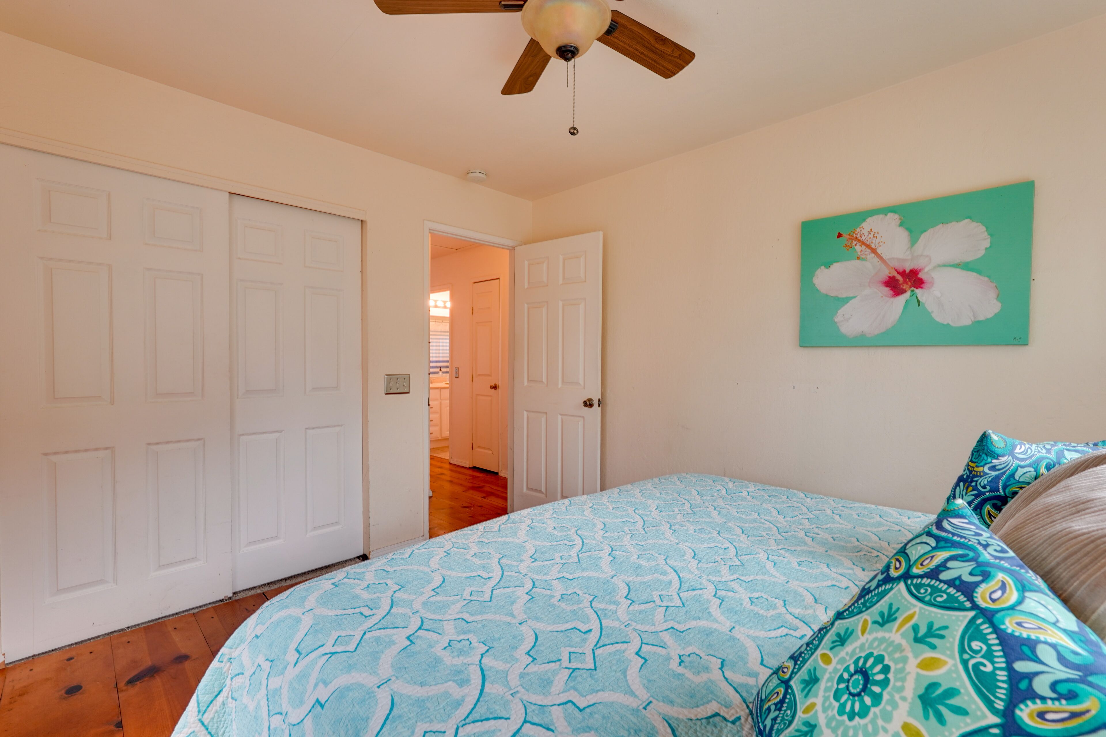 Un dormitorio con una cama, un ventilador de techo, una puerta y una pared con un cuadro floral.