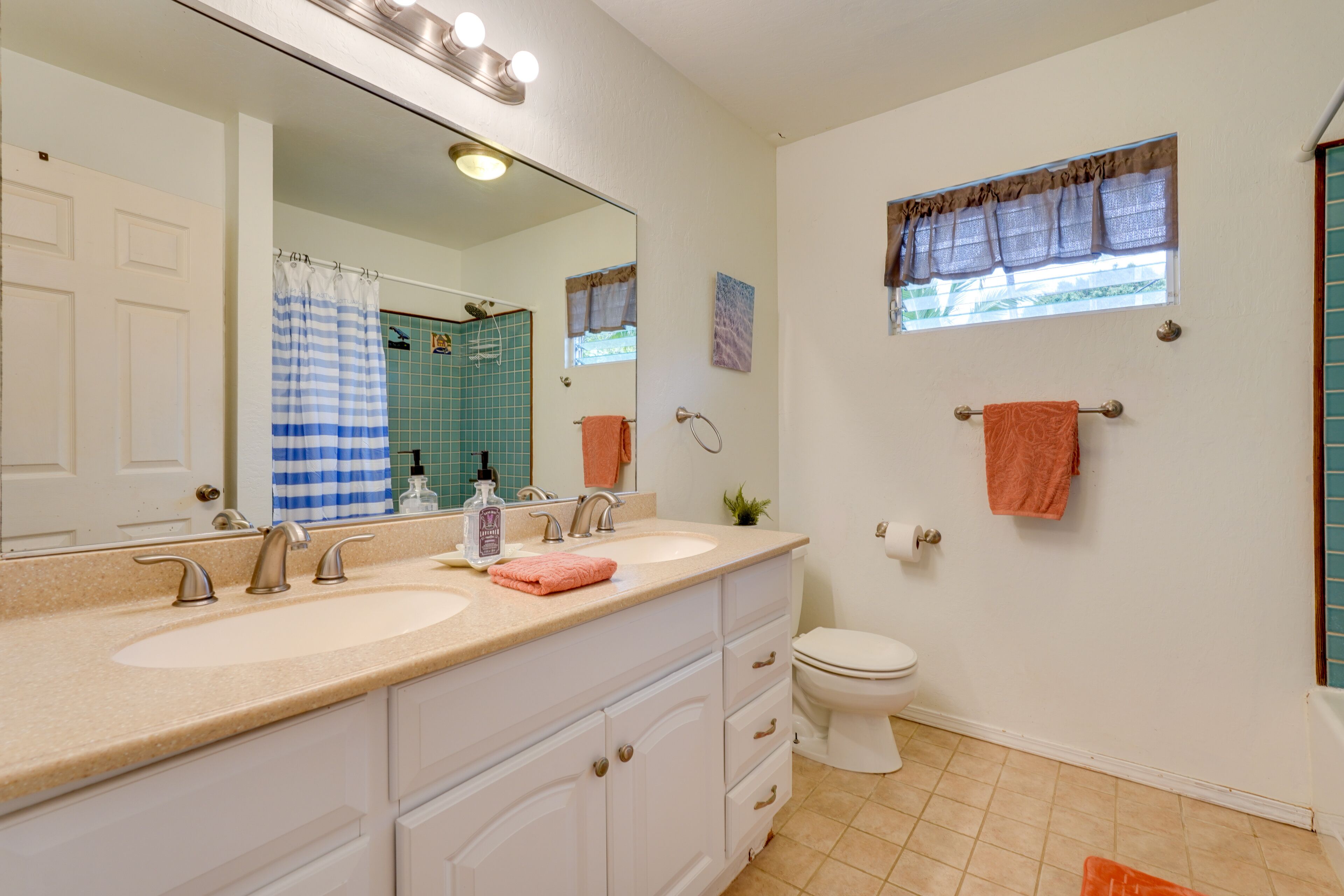 A bathroom with a double sink vanity, a toilet, and a window with a curtain.