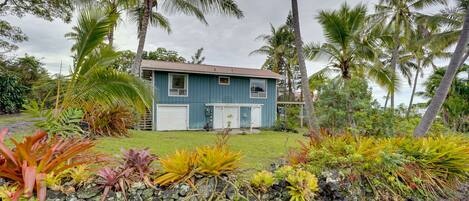 Une maison à deux étages, à l’extérieur bleu, entourée d’une végétation tropicale et de palmiers.