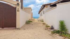 A narrow pathway leading to a beach, flanked by buildings with a sandy ground and some greenery.