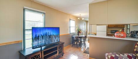 Uma sala de estar com televisão de tela plana, piso de madeira, área de jantar com mesa e cadeiras, e cozinha com armários e bancada.