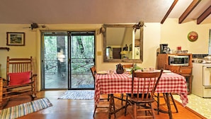Un comedor con mantel a cuadros rojo y blanco, sillas de madera, una mesa de comedor de madera, una mecedora de madera, un cojín rojo, piso de madera, un gran ventanal corredizo que da a un balcón, una cocina con microondas, estufa y armarios, y un espejo en la pared.